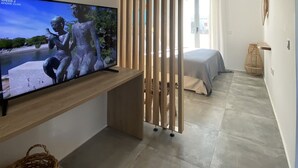Un dormitorio moderno con una televisión sobre un mueble de madera, una cama con cubrecama gris y un ventanal grande.