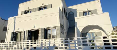 A modern two-story building with a white facade, multiple windows with shutters, and a white fence in the foreground.