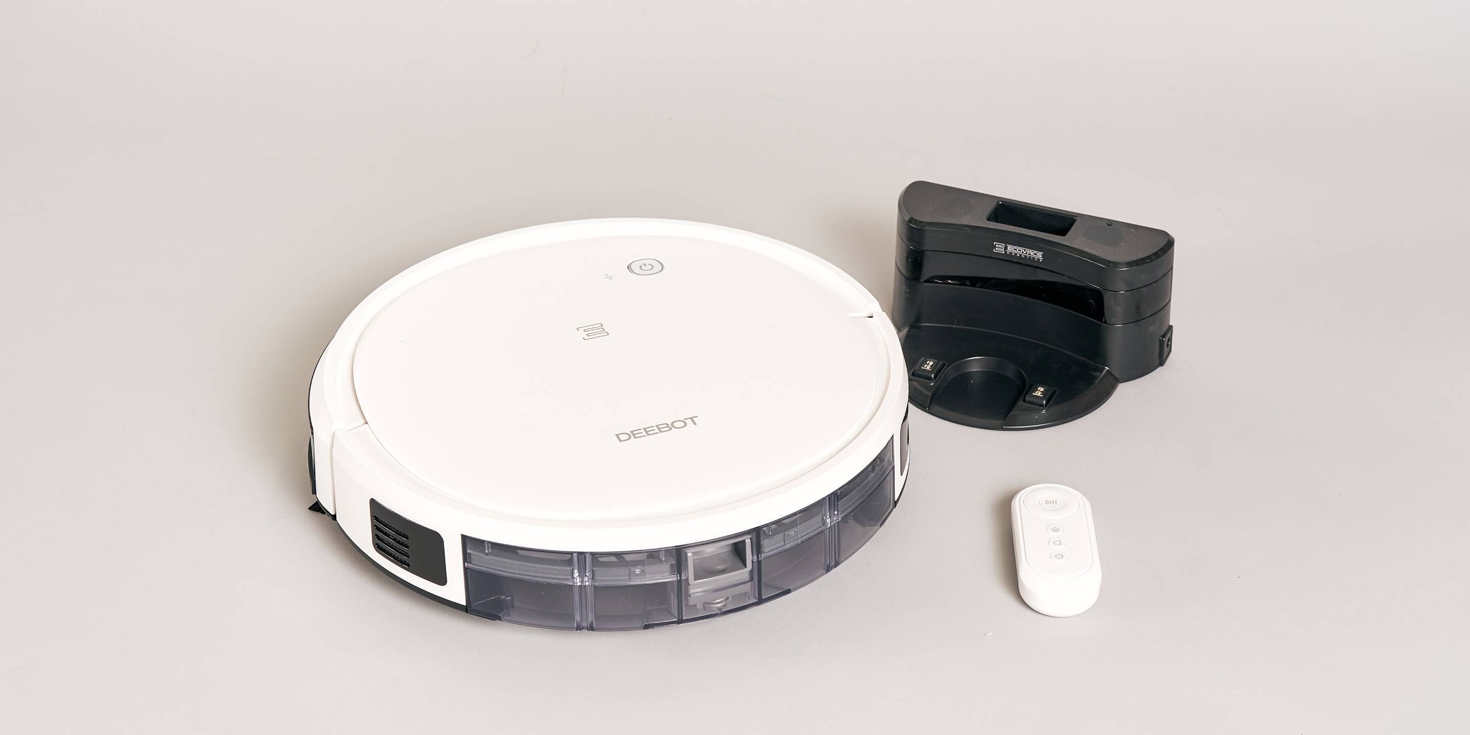 A white robotic vacuum cleaner with a transparent dustbin, a black charging dock, and a remote control.