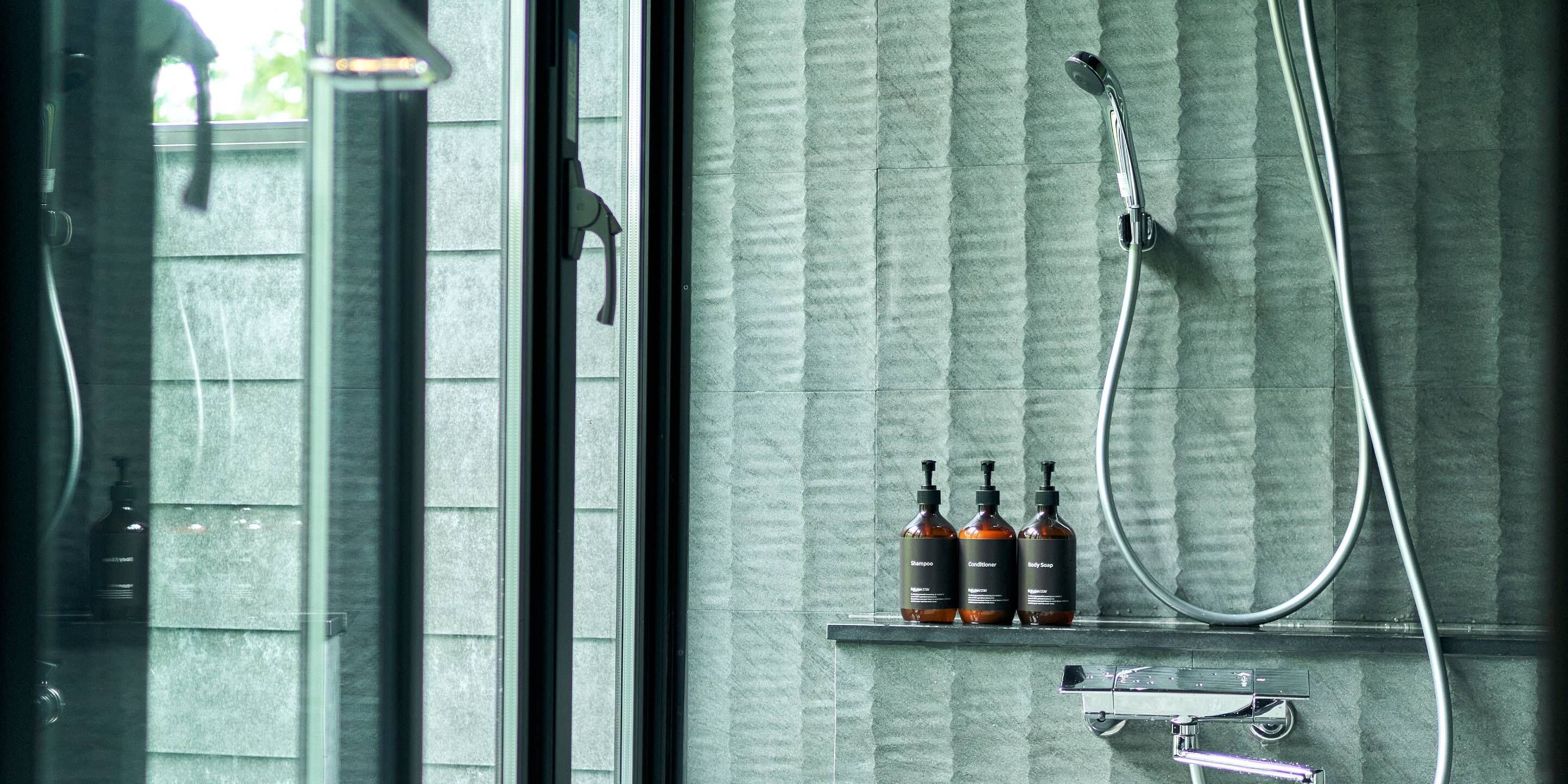 A bathroom with a shower, a glass door, and three bottles of shampoo, conditioner, and body wash on a shelf.