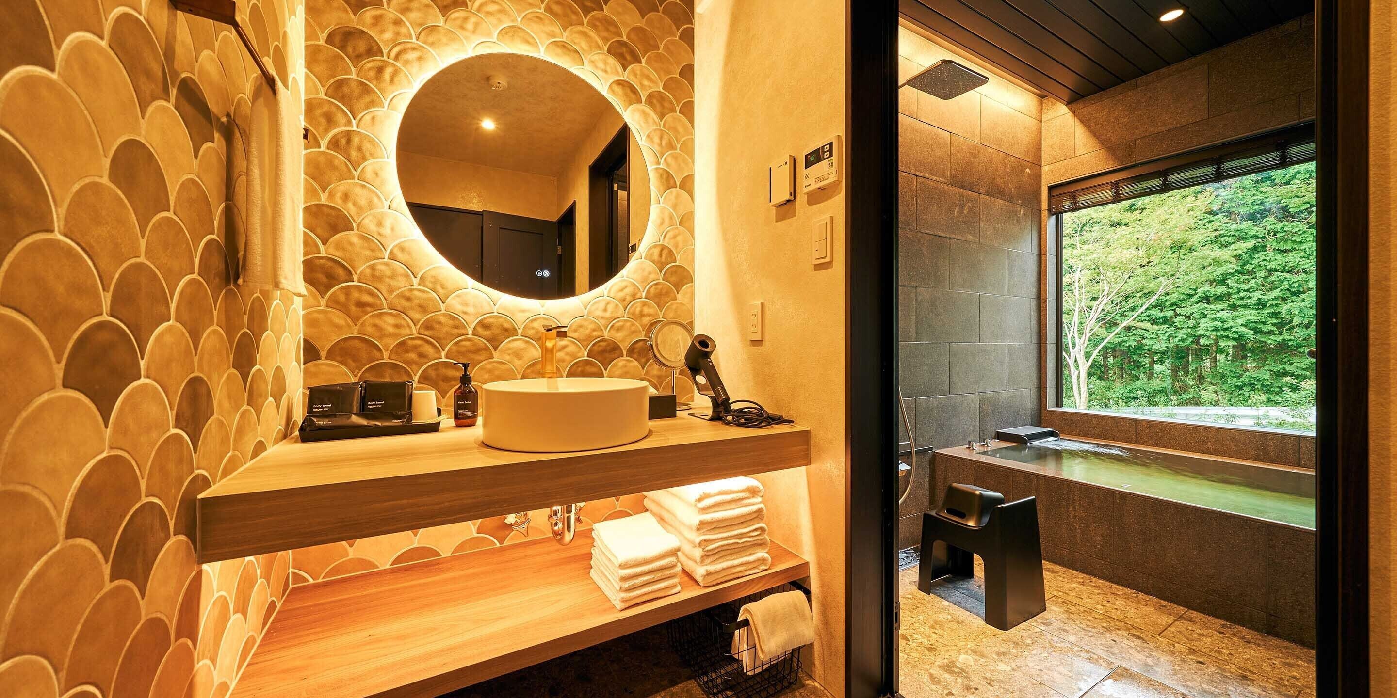 A modern bathroom with a round mirror, wooden vanity, and a view of greenery through a large window.