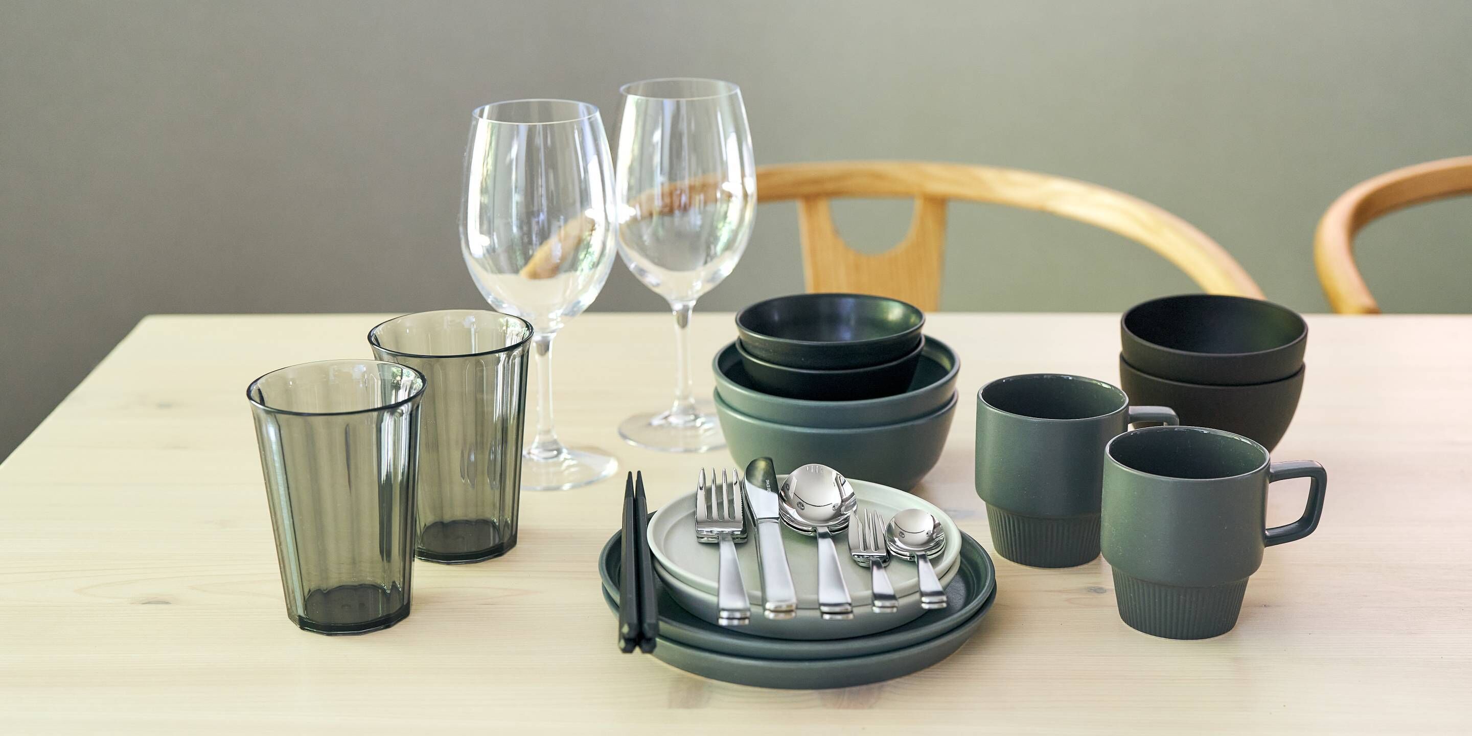 A dining table set with clear glasses, plates, cutlery, and dark-colored mugs.