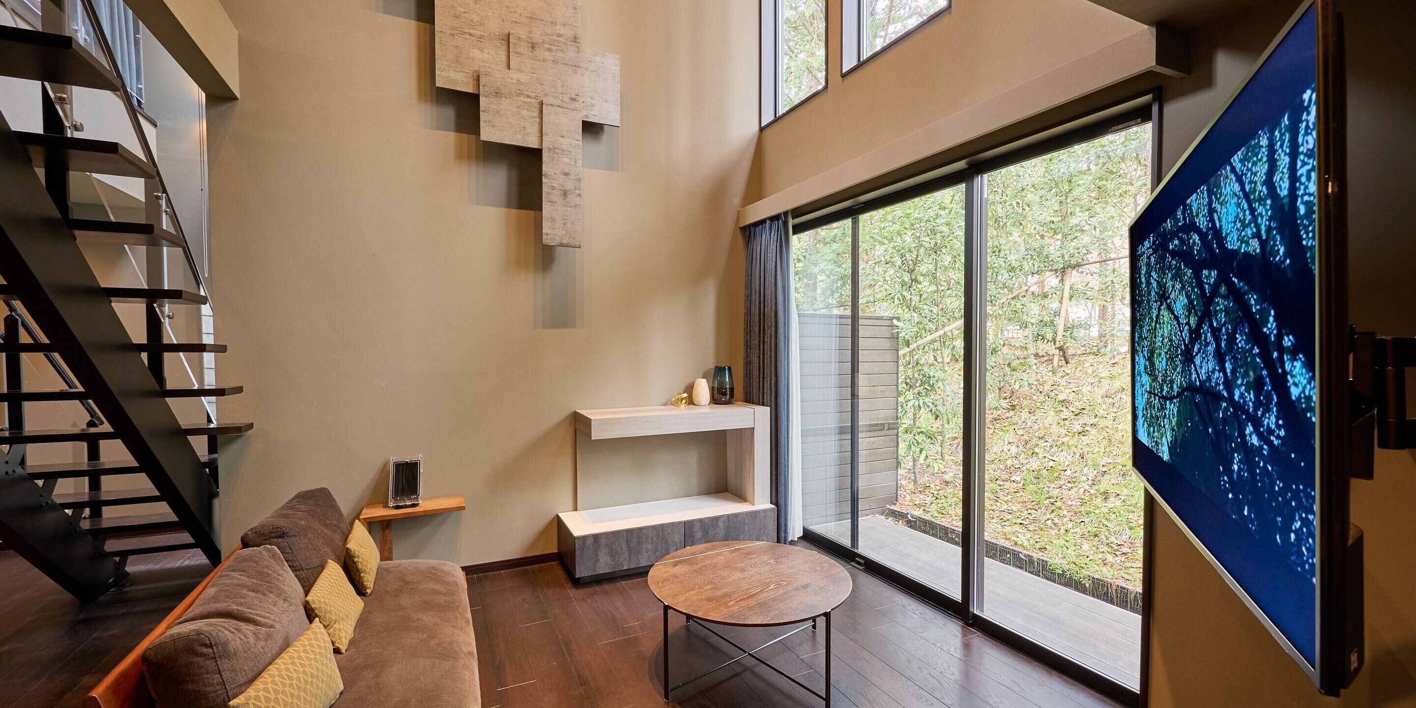 A modern living room with a staircase, a large TV, and a view of greenery outside.