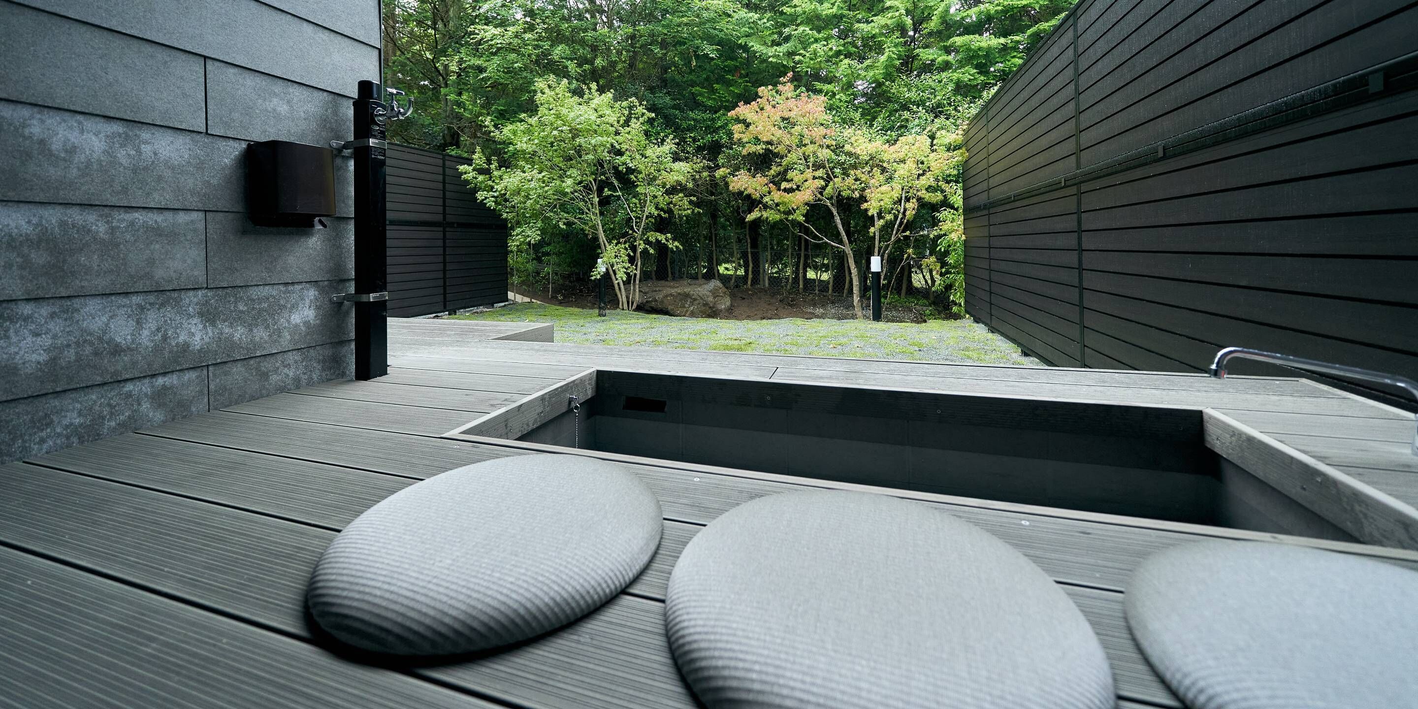 A modern outdoor deck with a built-in hot tub, seating area, and a view of a wooded area.