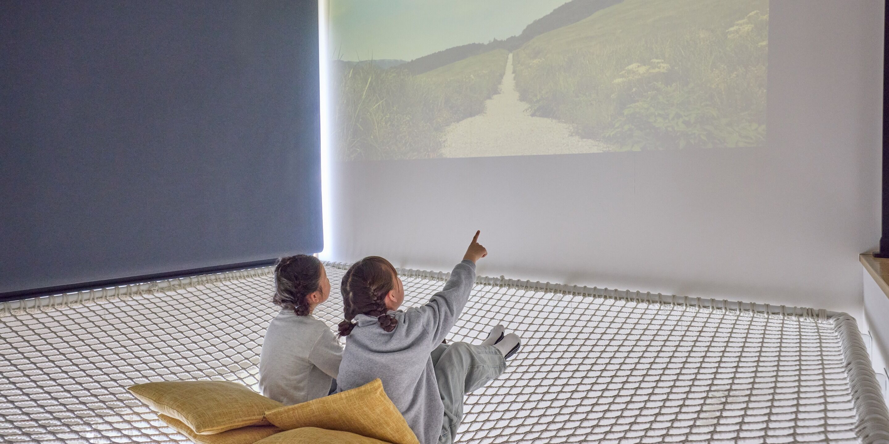 Two children sitting on a bed with a projection screen displaying a landscape.