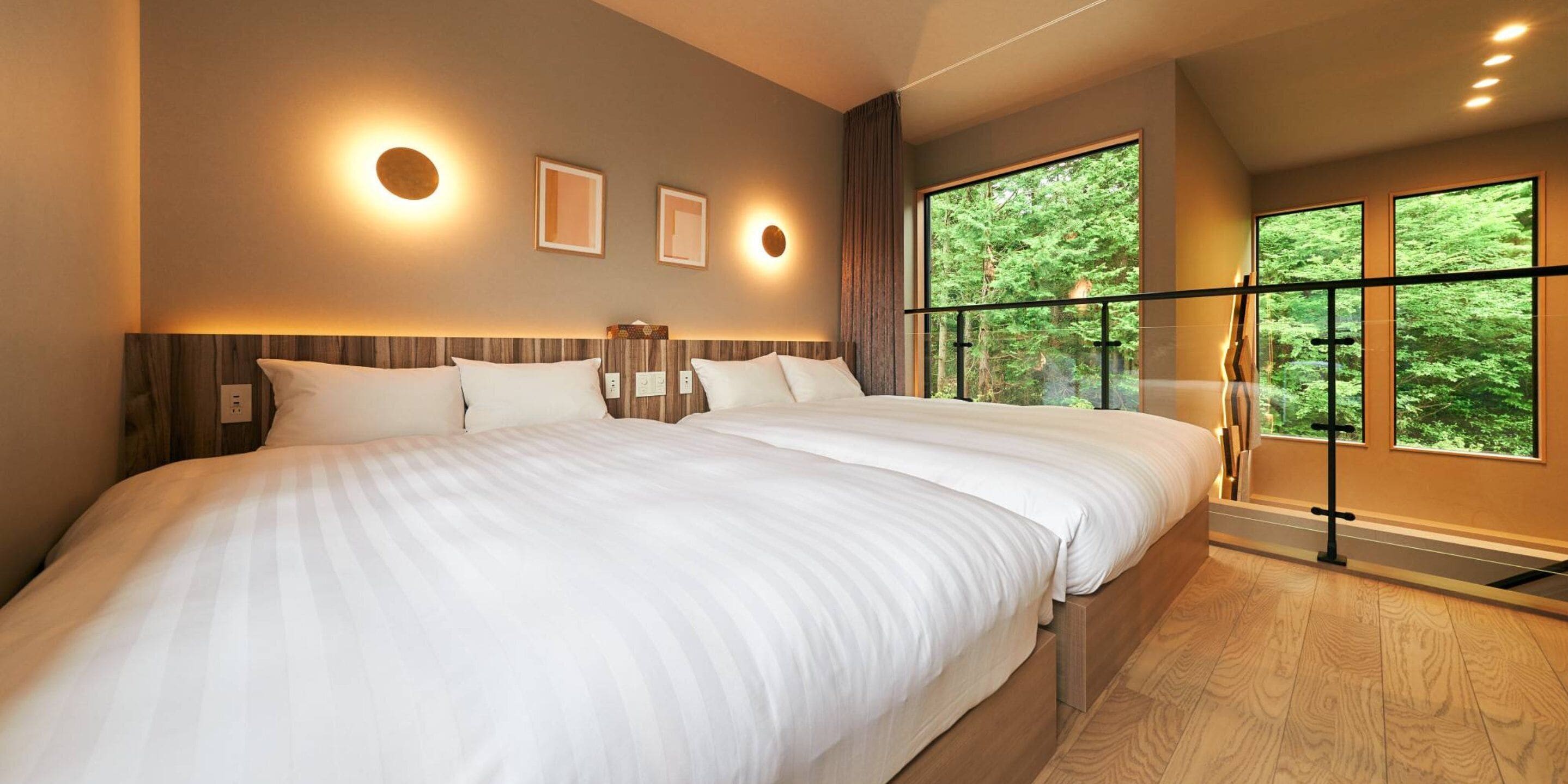 A modern hotel room with a large bed, a view of trees through a balcony, and wall-mounted lighting.