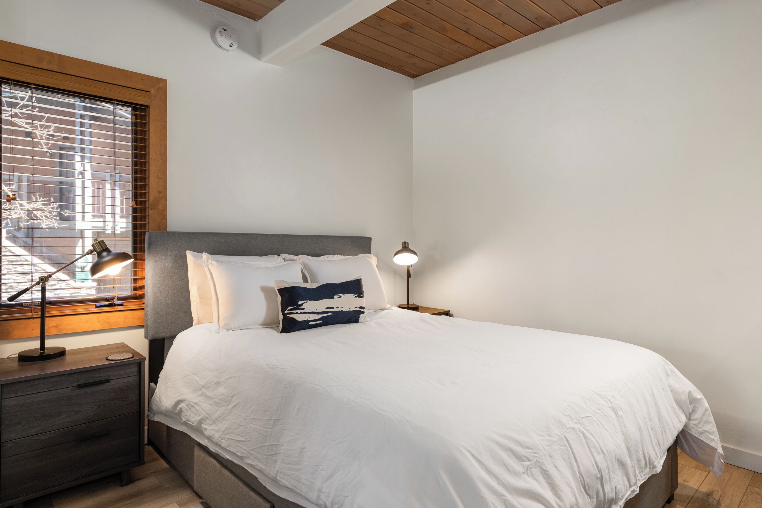 Quarto com cama, mesas de cabeceira, janela com persianas e teto com vigas de madeira.