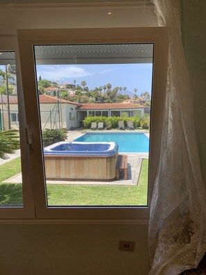 Uitzicht vanuit een raam met een houten jacuzzi, een zwembad en een gazon met stoelen.