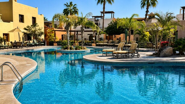Zona de piscina con sillones de descanso, palmeras y una piscina azul cristalina.