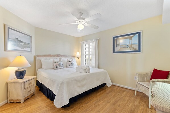 A bedroom with a bed, nightstand, chair, ceiling fan, and two framed pictures.