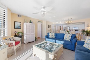 A living room with a blue sofa, a white armchair, a glass coffee table, and a ceiling fan.