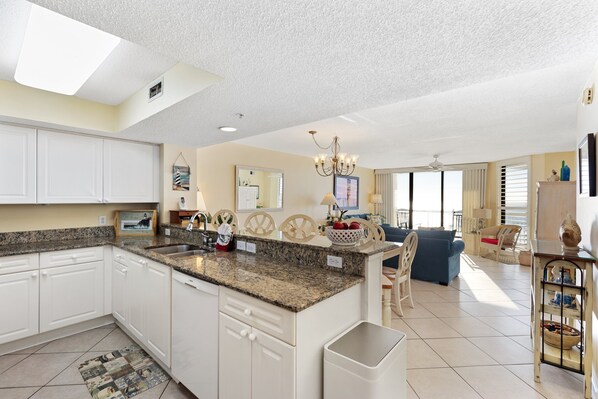 A spacious kitchen with granite countertops, white cabinetry, and a central island. There is a dining area with a blue sofa and a chandelier.
