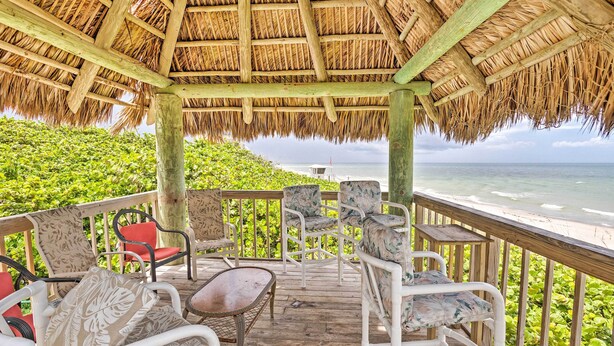 A covered outdoor seating area with wicker furniture and a thatched roof, overlooking a beach. - Jensen Beach Home: Walk to Beach Access! (Jensen Beach)