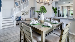 A dining area with a wooden table set for six, surrounded by chairs. A large plant is in the center, and a chandelier hangs above. - Windsor Hills: Sleeps 10, Pool, Spa, Game Room (Kissimmee)