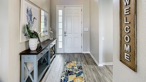 A hallway with a welcome sign, a white door, a console table with decorative items, and a floral rug. - Windsor Hills: Sleeps 10, Pool, Spa, Game Room (Kissimmee)