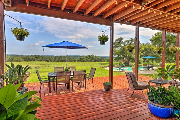 Una terraza de madera con mobiliario exterior, plantas colgantes y vistas a una piscina y paisaje verde.