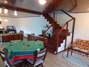 Uma sala de estar aconchegante com escada de madeira, uma mesa de jantar com toalha de mesa verde e uma televisão ao fundo.