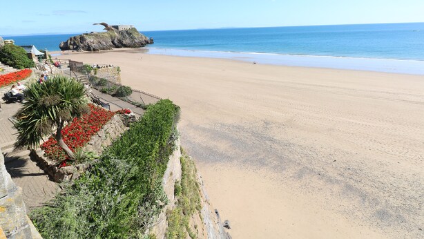 Uma praia costeira com um afloramento rochoso, um calçadão pavimentado e um muro de pedra com flores vermelhas.