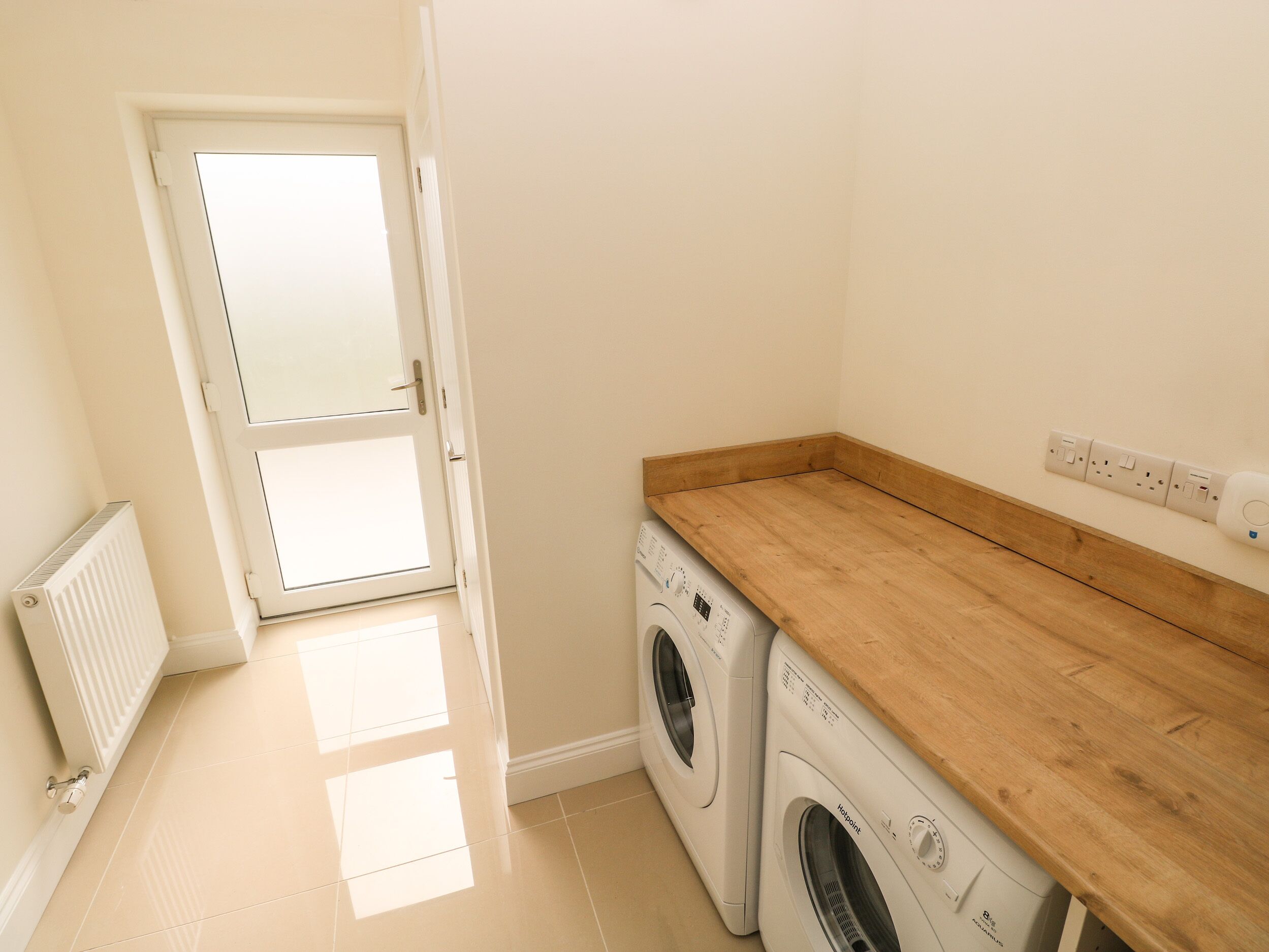 A laundry room with a wooden countertop, a washing machine, a dryer, a radiator, and a door leading to another room.