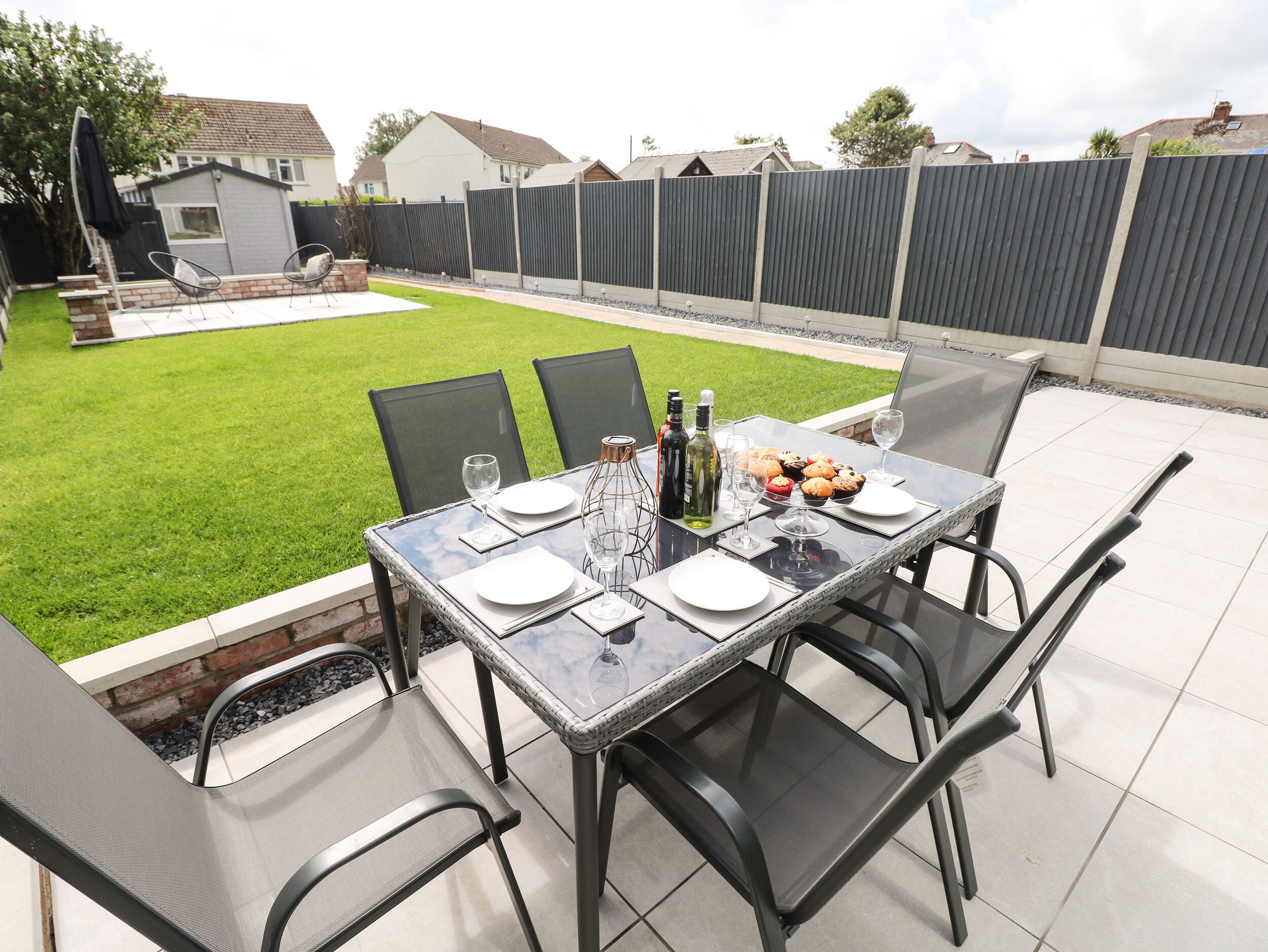 Eine Terrasse im Freien mit einem für vier Personen gedeckten Tisch, umgeben von Stühlen, mit Blick auf einen gepflegten Rasen und einen Zaun im Hintergrund.
