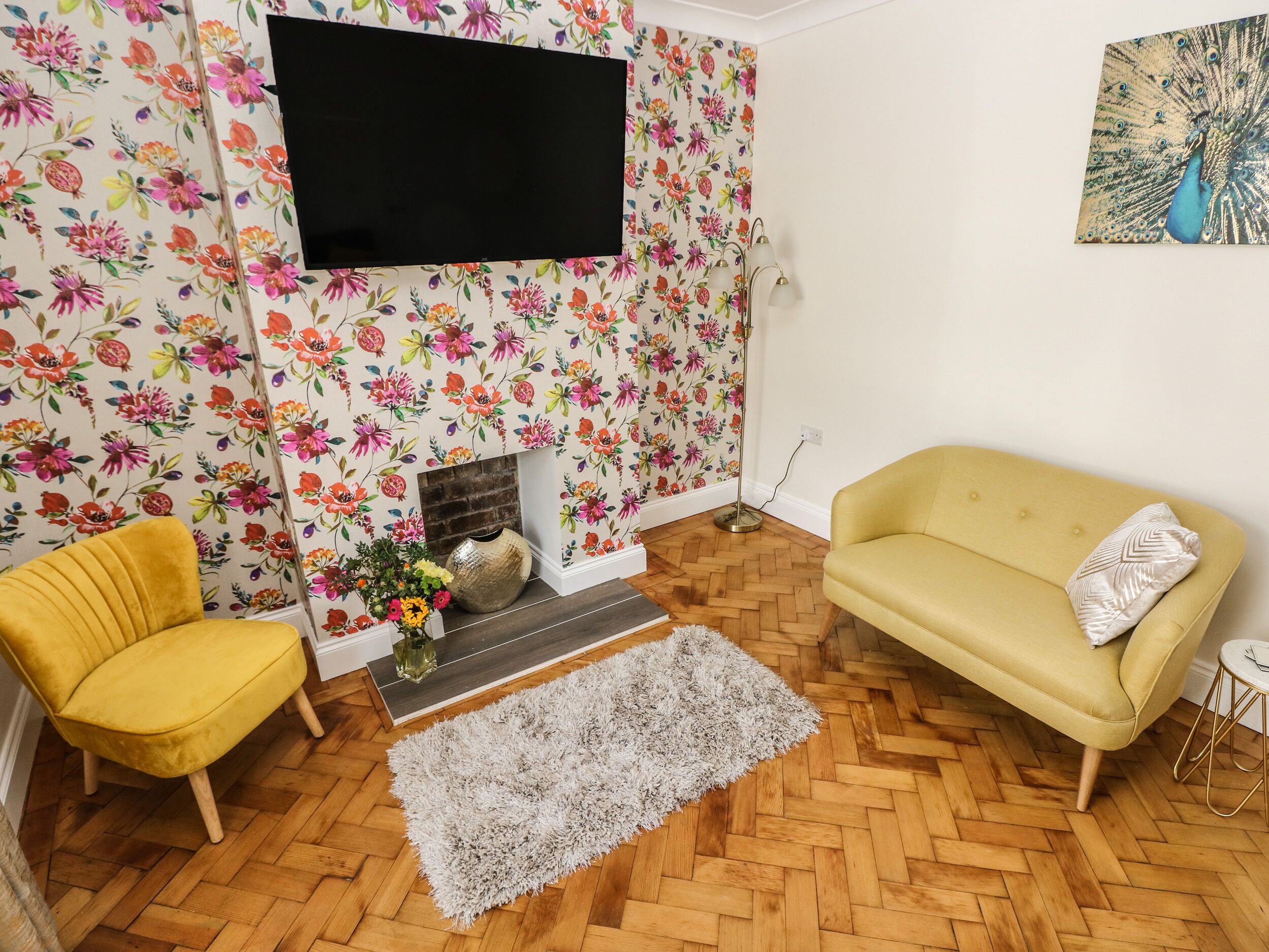 A living room with a floral wallpaper, a yellow sofa, a yellow armchair, a fireplace, and a flat-screen TV mounted on the wall.