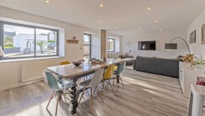 A spacious living area with a large dining table, colorful chairs, a sofa, and a TV mounted on the wall.