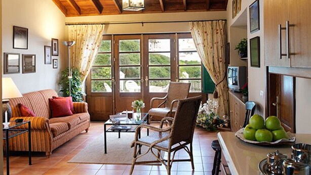 Ein gemütliches Wohnzimmer mit Holzboden, einer Couch, einem Couchtisch und einem Sessel. Es gibt Fenster mit Vorhängen, einen Fernseher und eine Küchenzeile mit Obstschale.