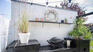 An outdoor kitchen area with a grill, potted plants, and a decorative wreath on a white wall.