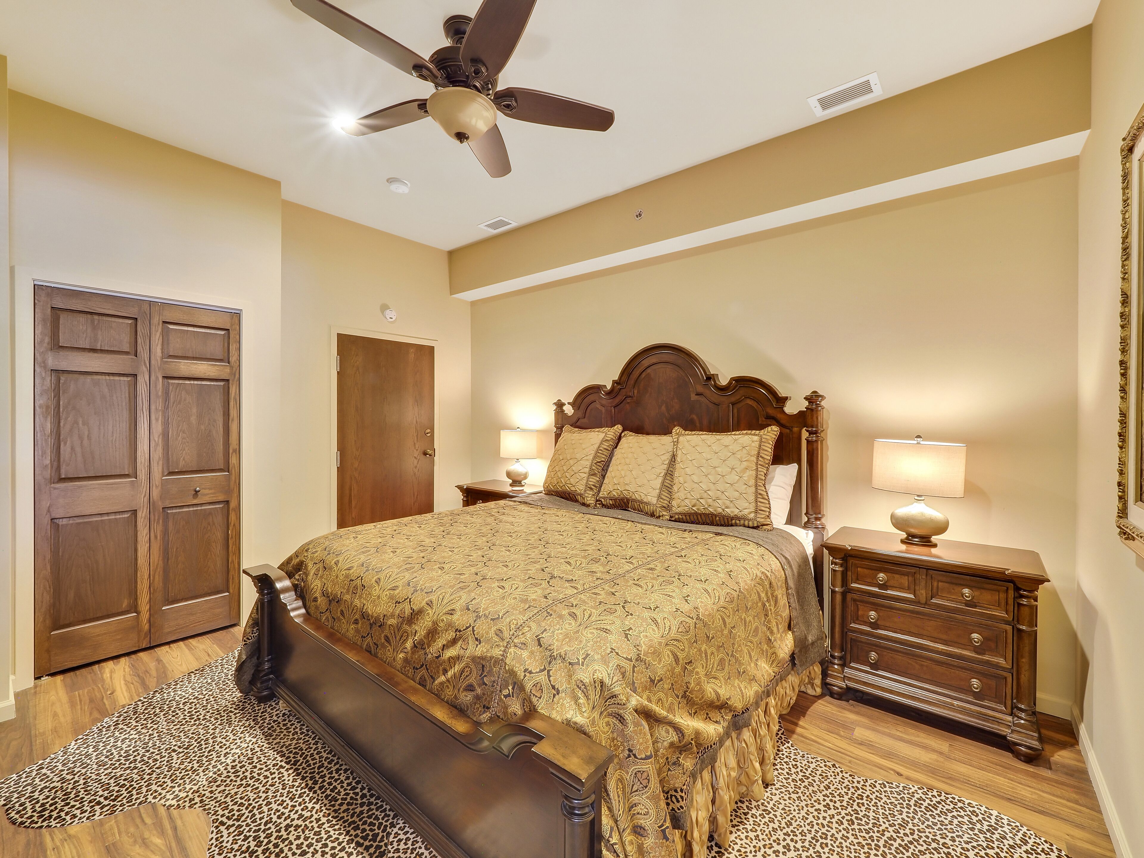 Une chambre avec un grand lit, un ventilateur de plafond, une commode en bois et un tapis à motifs léopard.