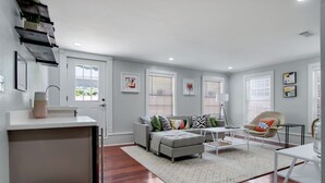 A modern living room with a grey sofa, a coffee table, and a chair. There is a white desk with a plant on it. - Canal DOWN |Streetcar Line MidCity| Add More Space (New Orleans)
