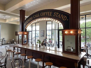 En kaffevogn med et skilt, der siger "Morning Call Coffee Stand", i en café med barstole og borde.