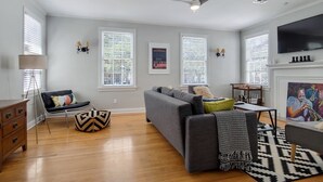 Ein Wohnzimmer mit grauem Sofa, einer Holzkommode, einer Stehlampe und einem schwarz-weißen Teppich.