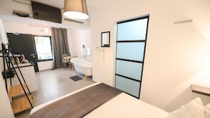 A modern hotel room with a bed, a desk with a chair, a bathroom with a bathtub, and a large window.