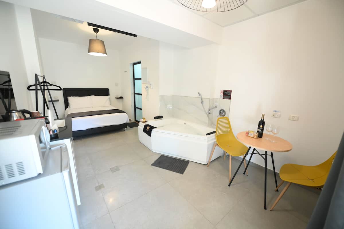 A modern hotel room with a bed, a bathtub, a desk with a computer, and a small dining table with yellow chairs.