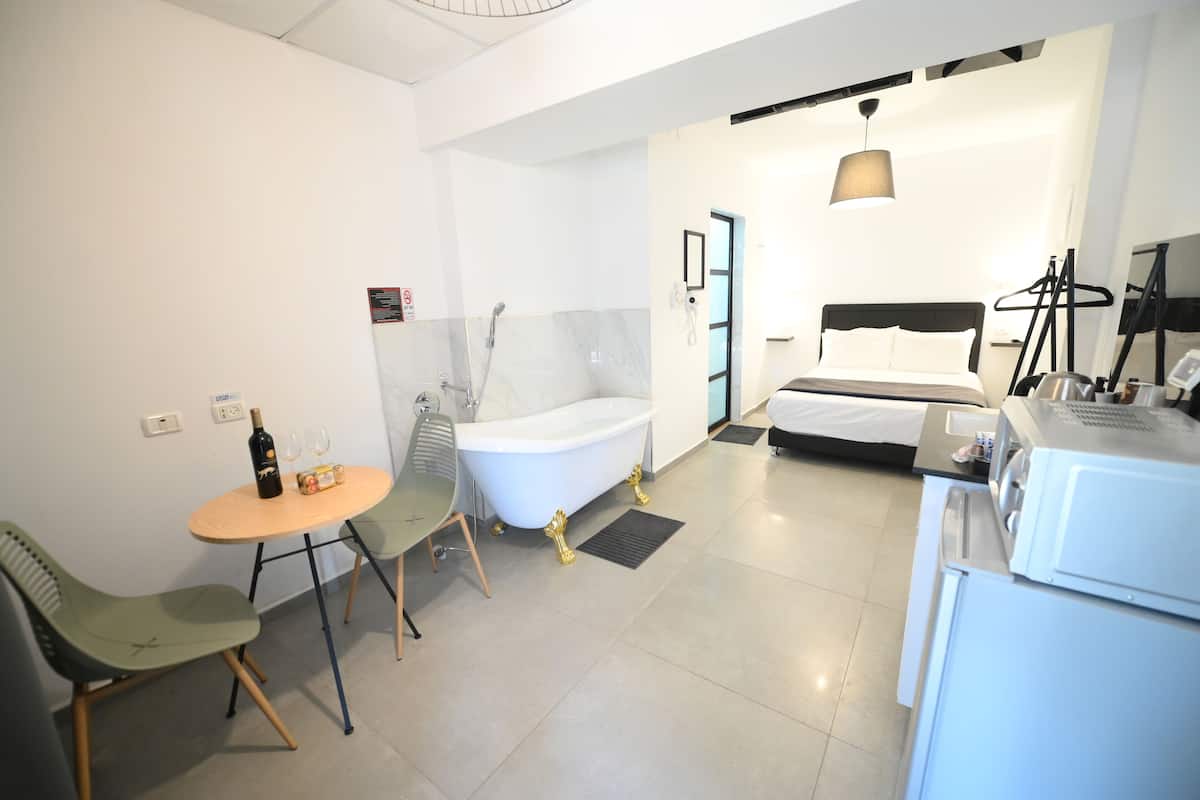 A modern hotel room with a bathtub, a bed, a dining area with chairs, and a small table with a bottle and glasses.