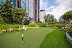A miniature golf course with a green artificial turf, surrounded by lush greenery and modern buildings in the background.