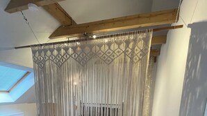 Une chambre avec un lit placé sous un dais en macramé, des poutres en bois au plafond et une lucarne.