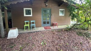 Une petite cabane en bois, avec un toit en tôle ondulée, deux fenêtres et une porte. On y trouve deux chaises et une petite table à l’extérieur.