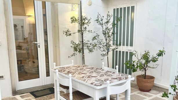 Un espace patio avec une table et des chaises blanches, des plantes en pot et une porte-fenêtre coulissante donnant sur un espace intérieur.