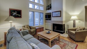 Een ruime woonkamer met een grote bank, een salontafel en twee fauteuils. Er is een open haard, een televisie en een groot raam dat zorgt voor daglicht.