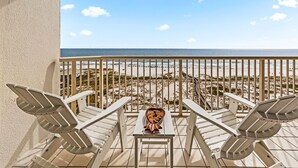 Un balcon avec deux fauteuils de jardin blancs et une petite table agrémentée d’un objet décoratif, donnant sur une plage avec une rambarde et l’océan en arrière-plan.