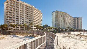 Un hôtel situé en bord de mer, avec une promenade en bois menant à la plage, entouré de palmiers et surplombé d’un ciel bleu azur.