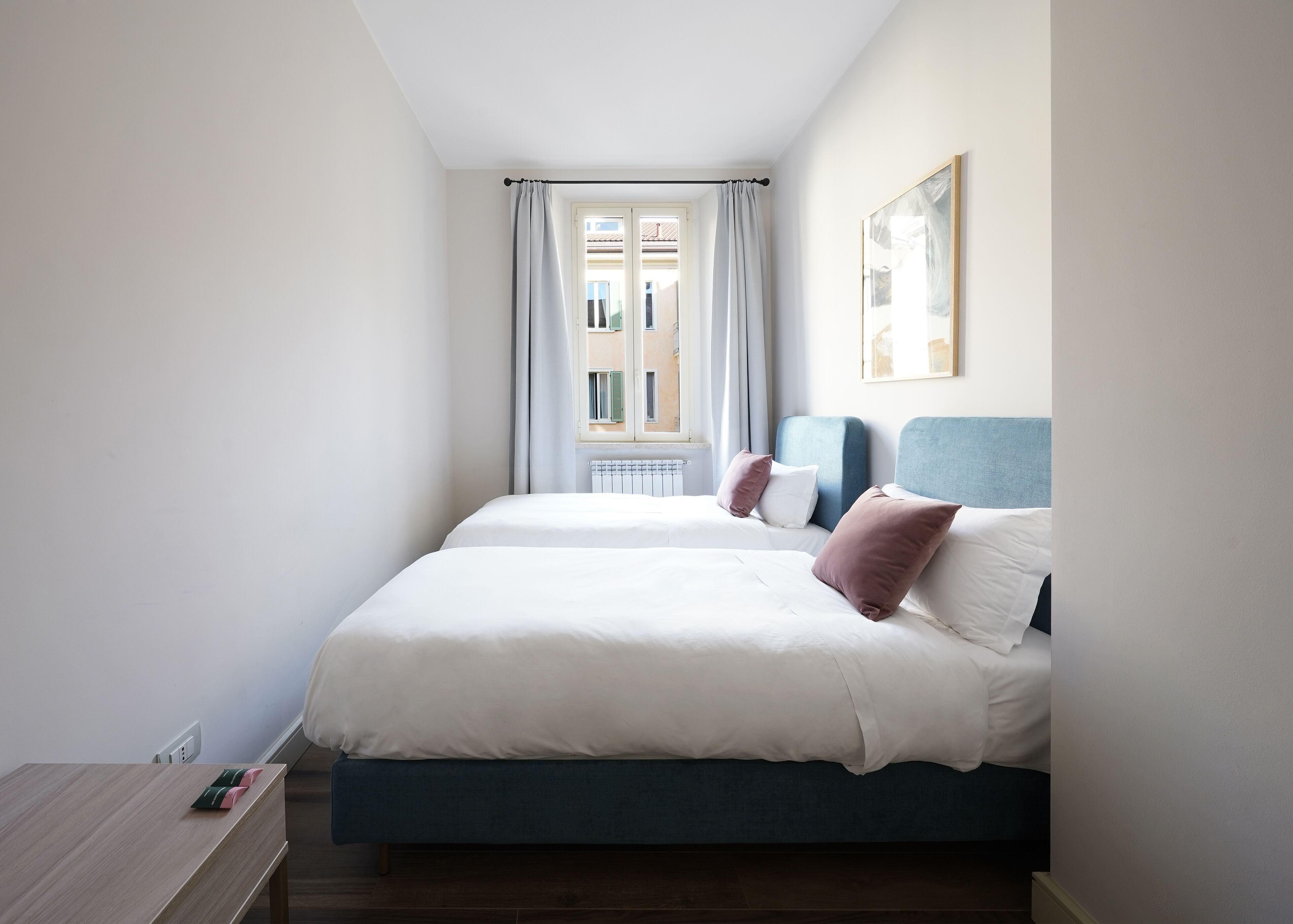 A hotel room with two beds, a window with curtains, a radiator, and a framed picture on the wall.
