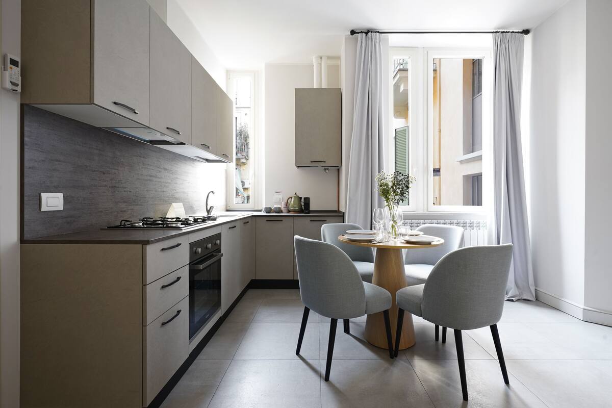 Una cocina moderna con comedor, que incluye una isla central, sillas grises y un ventanal con cortinas.