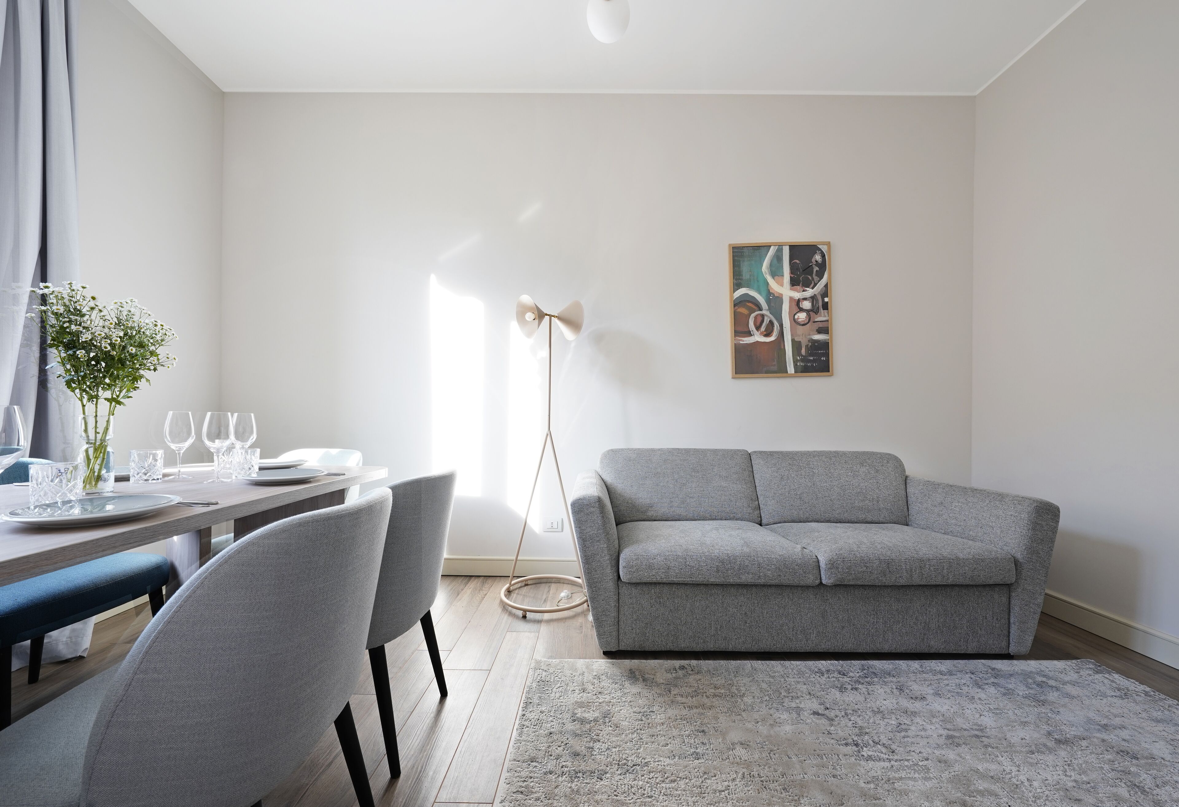 A modern living room with a grey sofa, a dining table set with plates and glasses, a hanging floor lamp, and a framed abstract painting on the wall.