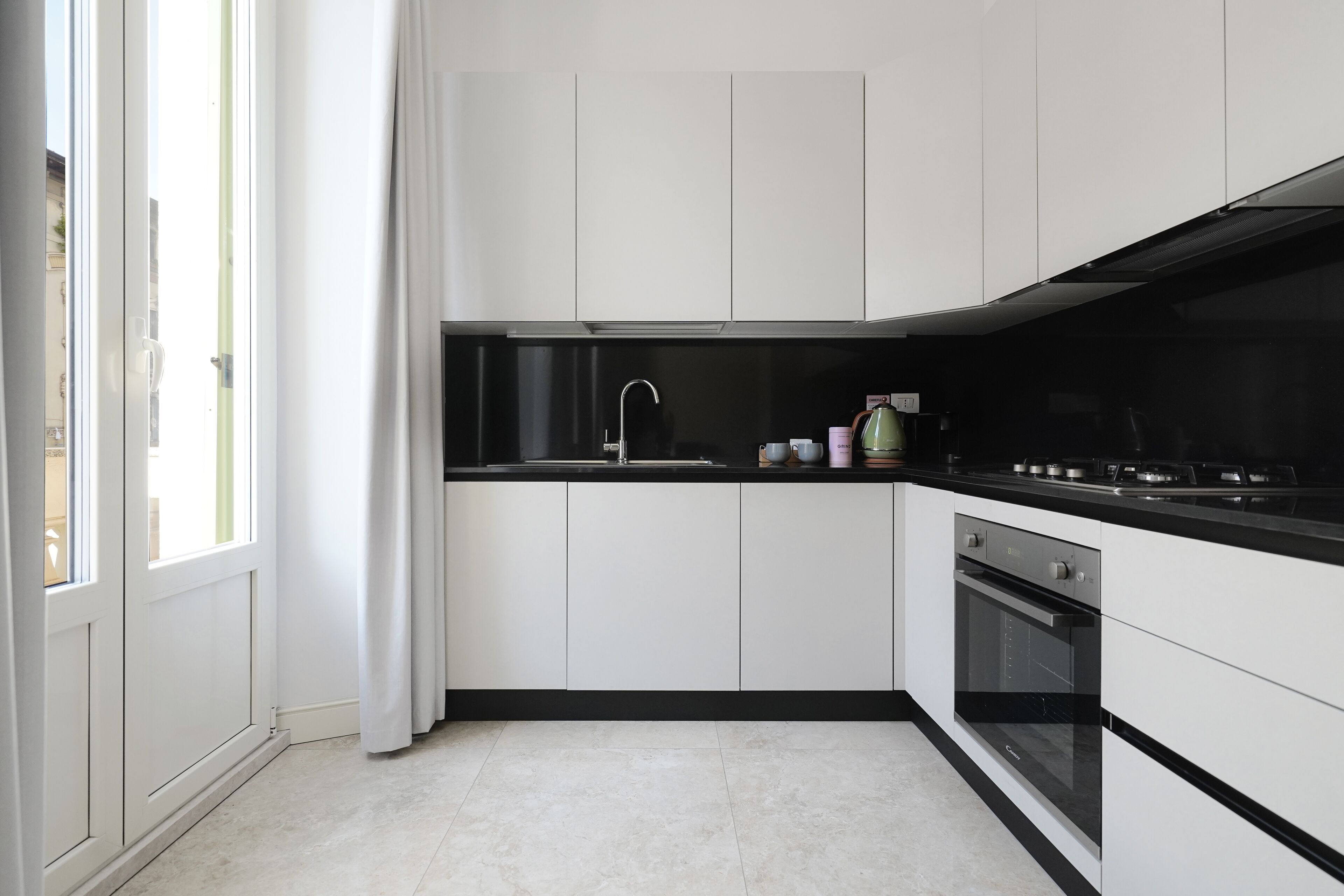 Une cuisine moderne avec des meubles blancs, un plan de travail noir, un four encastré et une fenêtre agrémentée de rideaux blancs.