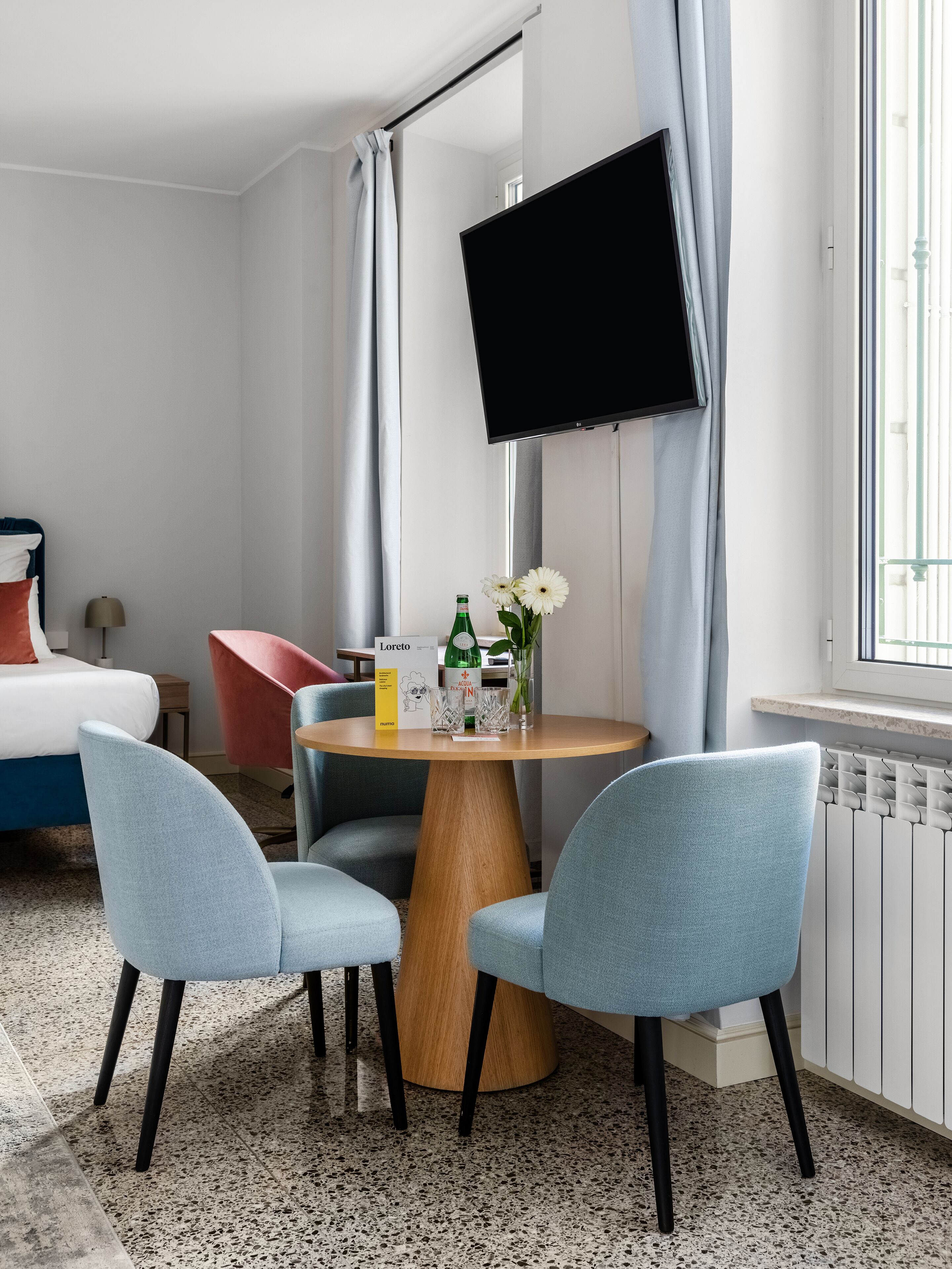 A hotel room with a bed, a desk with a chair, a TV mounted on the wall, and a window with curtains.
