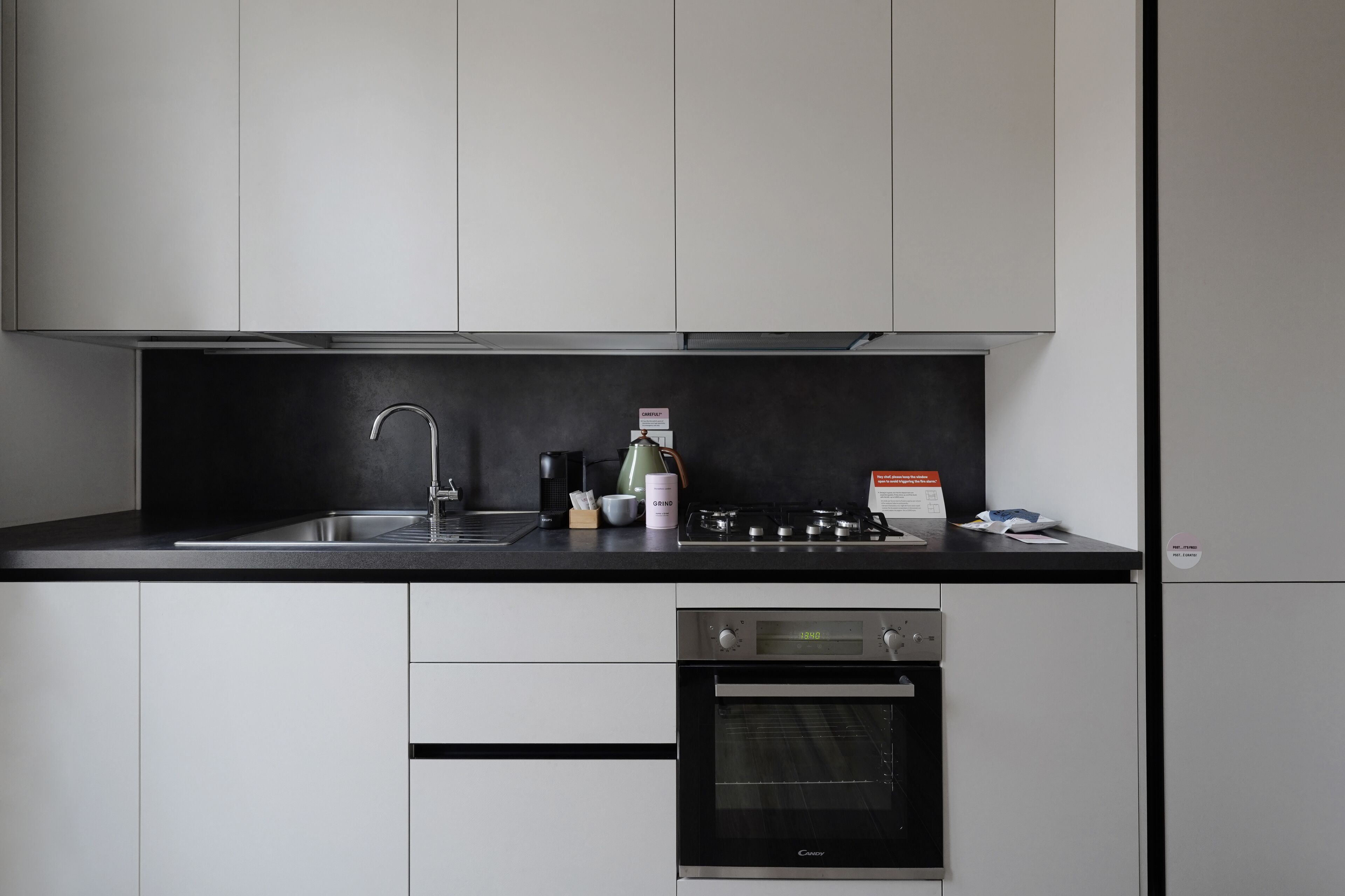 A modern kitchen with white cabinets, a black countertop, a stainless steel sink, a coffee maker, a kettle, and a built-in oven.