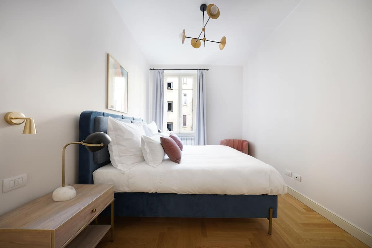 Un dormitorio con cama, mesita de noche, lámpara y ventana con cortinas.