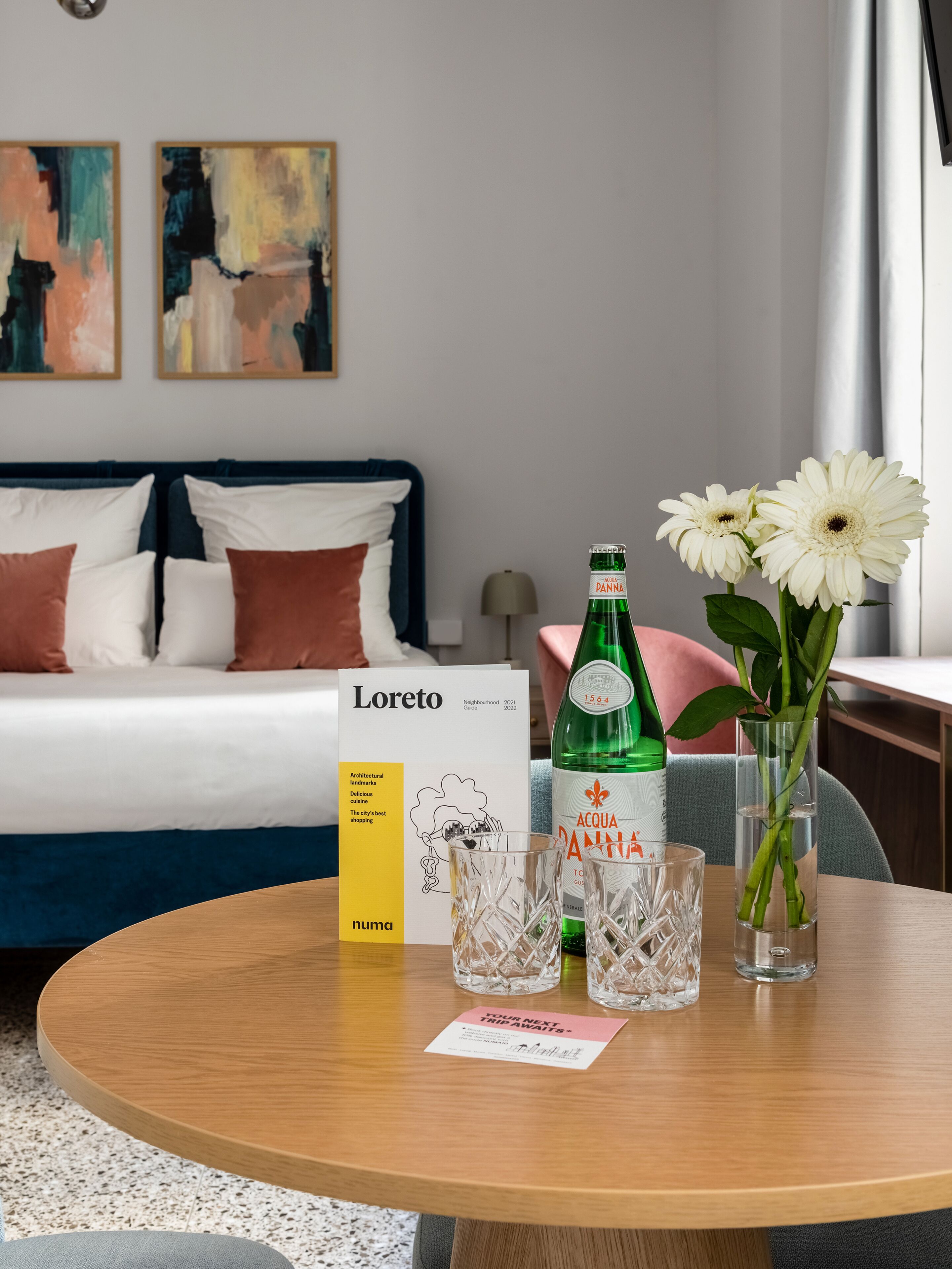 A hotel room with a bed, a table with a bottle of Acqua Panna, a glass, and a vase with flowers. There is a guidebook on the table.
